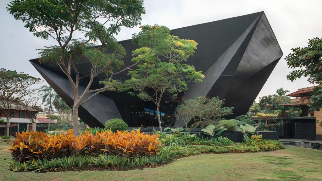A modern, angular black building with a faceted exterior stands amidst lush green trees and landscaped gardens. Vibrant orange and yellow flowers contrast beautifully with the dark structure, all meticulously approved through HOA architectural reviews to maintain community standards.