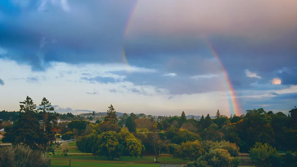 A scenic view of a lush green landscape beneath a cloudy sky with a visible rainbow arching in the distance. Dense trees cover most of the land, and a small path runs through the greenery. Seasonal maintenance keeps this area vibrant, as hills rise on the horizon.