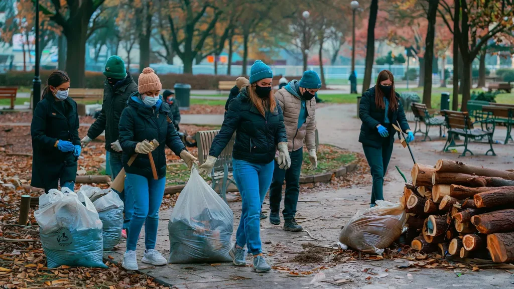 A group of people wearing masks and gloves perform seasonal maintenance in a park, picking up trash and debris. Fallen leaves and logs are scattered on the ground as colorful autumn trees surround them, ensuring the Bay Area HOAs keep spaces clean and enjoyable for all.