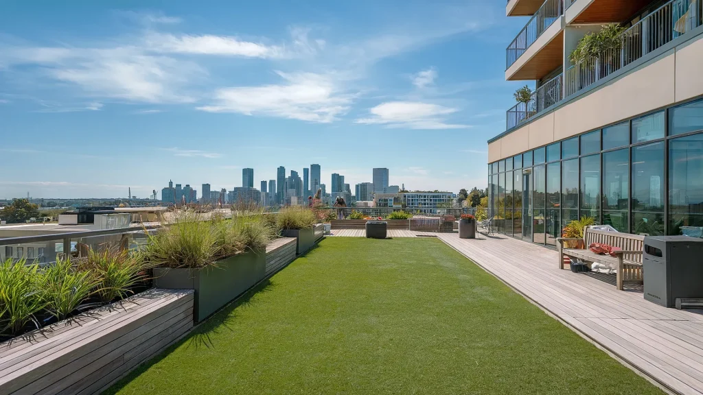 A rooftop with a lawn area and planters offers a serene escape, overlooking a city skyline against a clear blue sky. The building on the right boasts large glass windows and a balcony adorned with potted plants, reflecting East Bay property value trends.