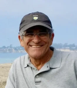 A smiling older man wearing glasses, a black cap, and a gray polo shirt stands outdoors with a blurred background of water and distant land, reflecting the approachable spirit of an experienced Oakland HOA Management team.