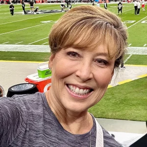 A woman with short, light brown hair and a gray shirt smiles for a selfie at a football stadium, with the field, sideline, and other people visible in the background—capturing the fun spirit of an Oakland HOA management team outing.