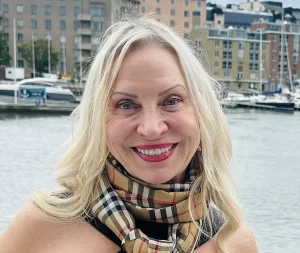 A smiling woman with long blonde hair stands in front of a waterfront with boats and modern apartment buildings—perhaps the ideal spot for an Oakland HOA management team to oversee. She is wearing a beige, plaid scarf and a black top.