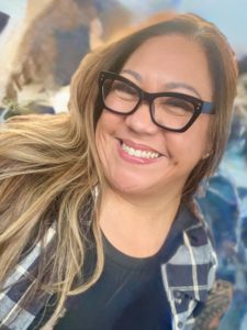 Suzie Garcia, a woman with long, light brown hair wearing black glasses, a black t-shirt, and a plaid shirt, smiles brightly at the camera. The background is softly blurred.
