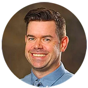 A man with short dark hair smiles at the camera, wearing a blue collared shirt and a patterned tie, against a neutral brown background.