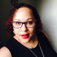 A woman with curly hair, red lipstick, and glasses is wearing a sleeveless black top and a necklace, posing stylishly as if ready for a Chanel resale in front of a light-colored wall.